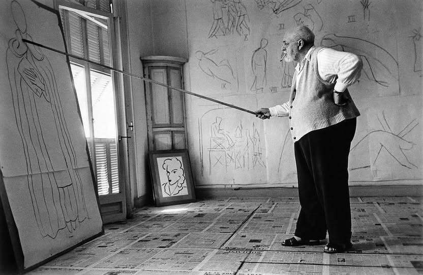 Matisse is standing in his studio drawing a mural with a very long bamboo stick. 