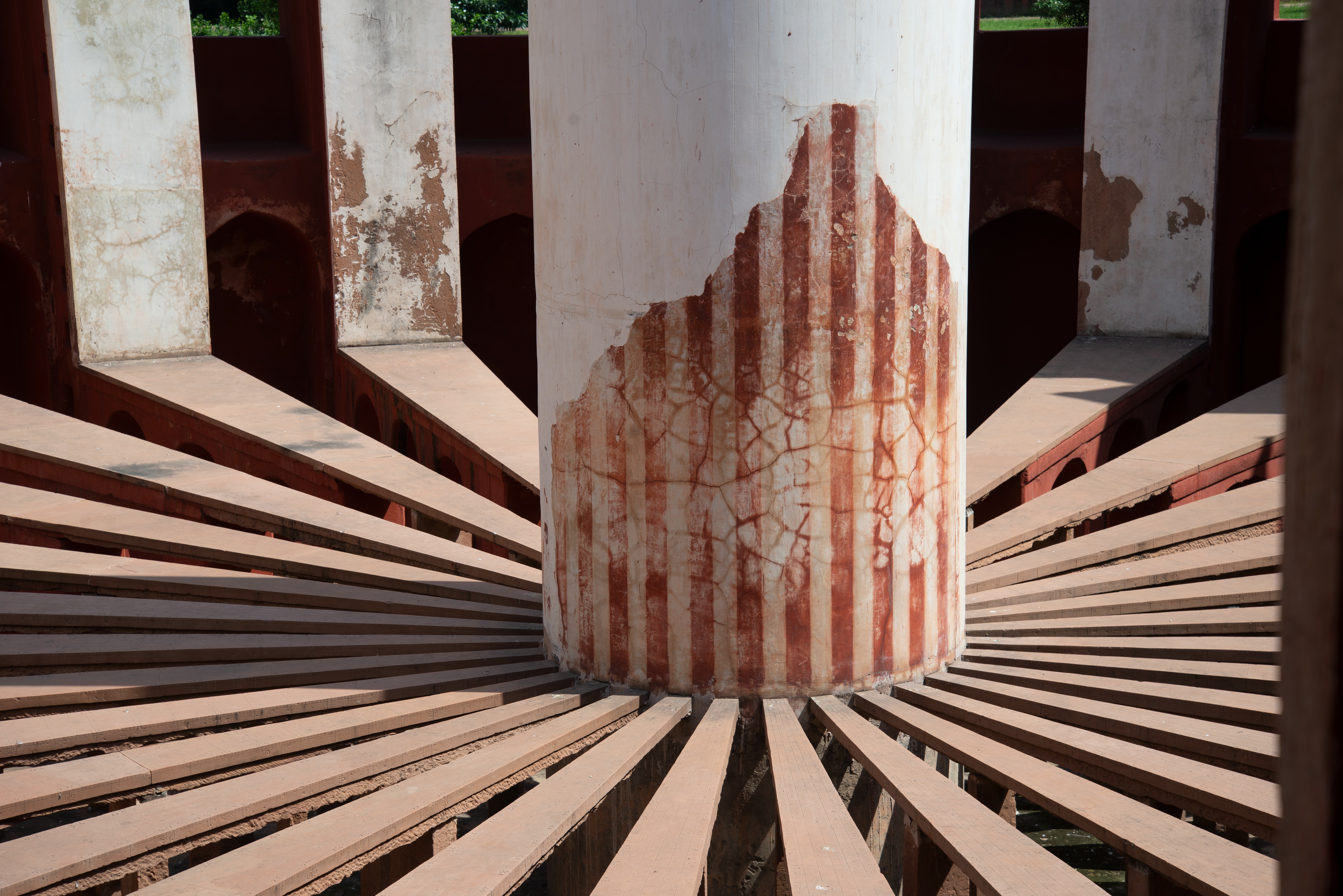 A large, weathered pillar is surrounded by a series of evenly spaced radial beams in a circular architectural structure. The structure is made of stone. 