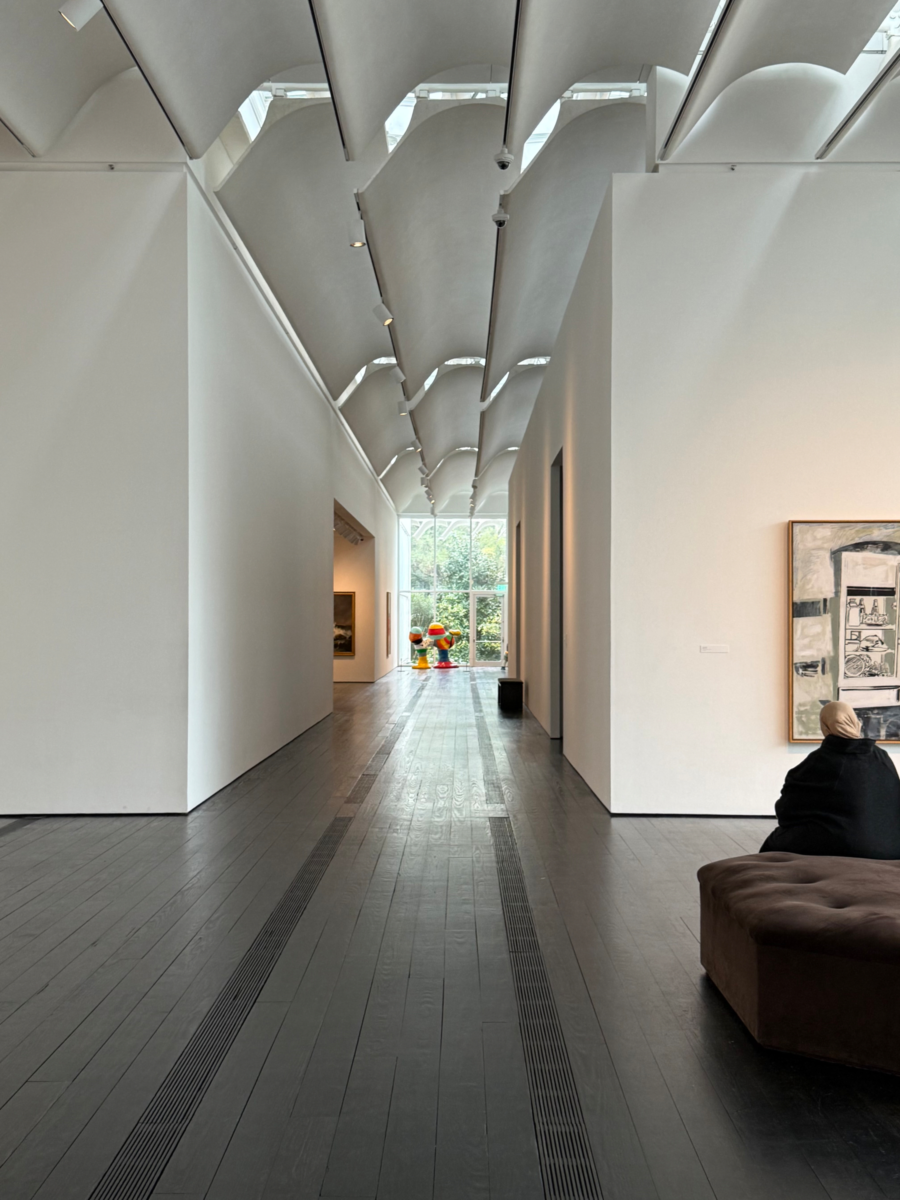 A modern art gallery features minimalist architecture with high ceilings and a walkway leading to a large window, where a person sits on a couch in the foreground. The buidling is the Menil Collection main building by Renzo Piano. 