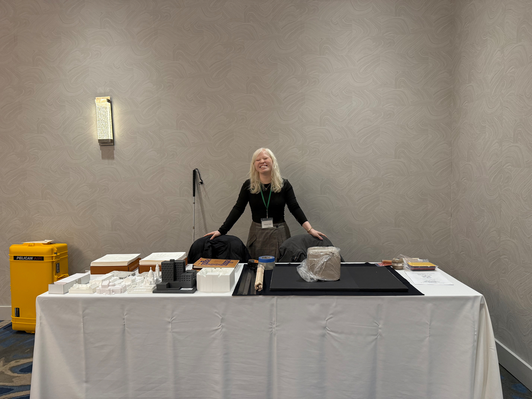 A photograph of Hannah, a Chinese-American woman with Albinism wearing all black and standing at a tactile table. She is gesturing with her arms towards a set of tactile architecture models and a lump of unsculpted air-dry clay for a co-designed architecture model we will be making tomorrow. 