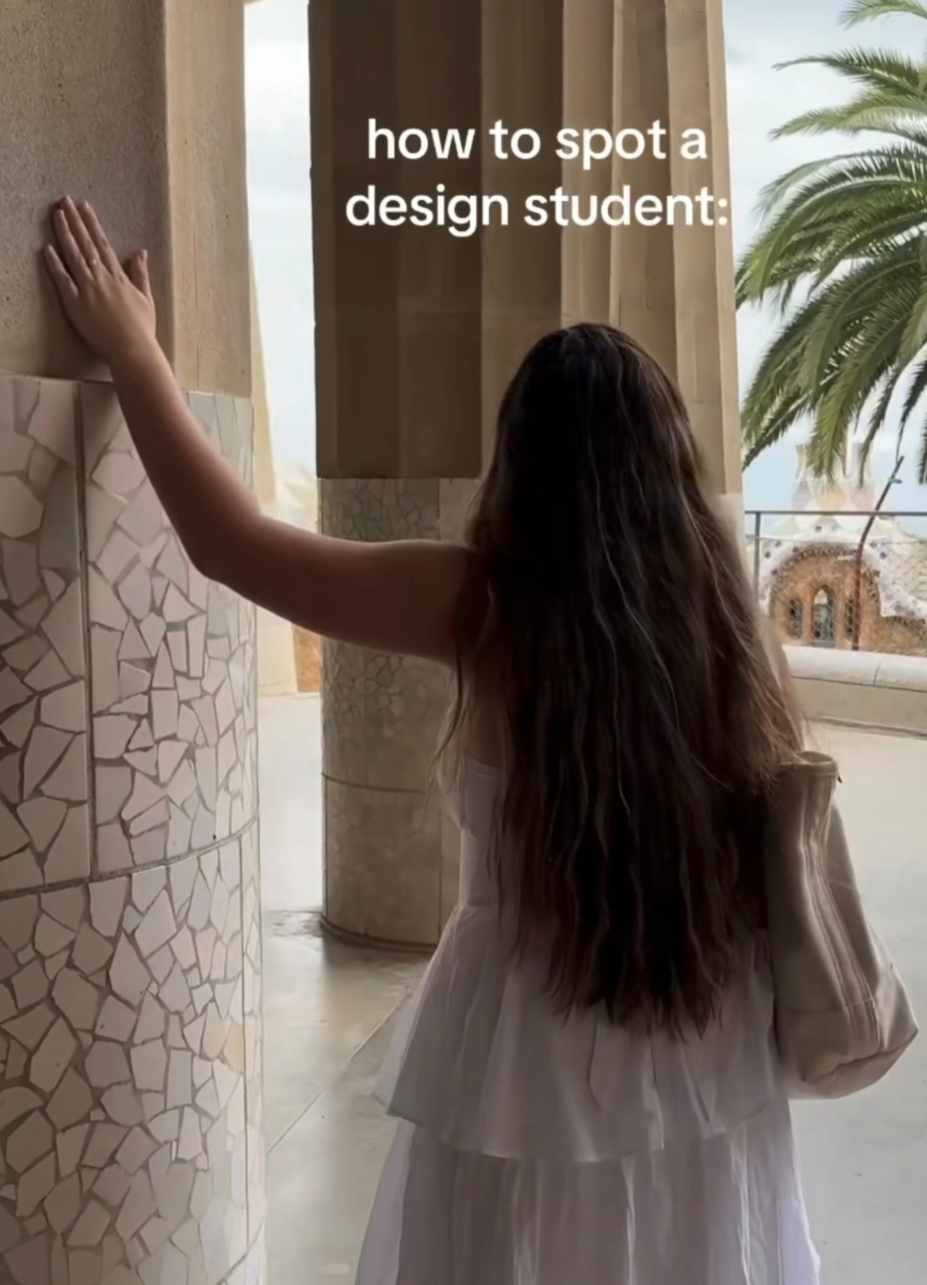 A person with long hair stands outdoors, touching a textured wall, with a palm tree visible in the background.