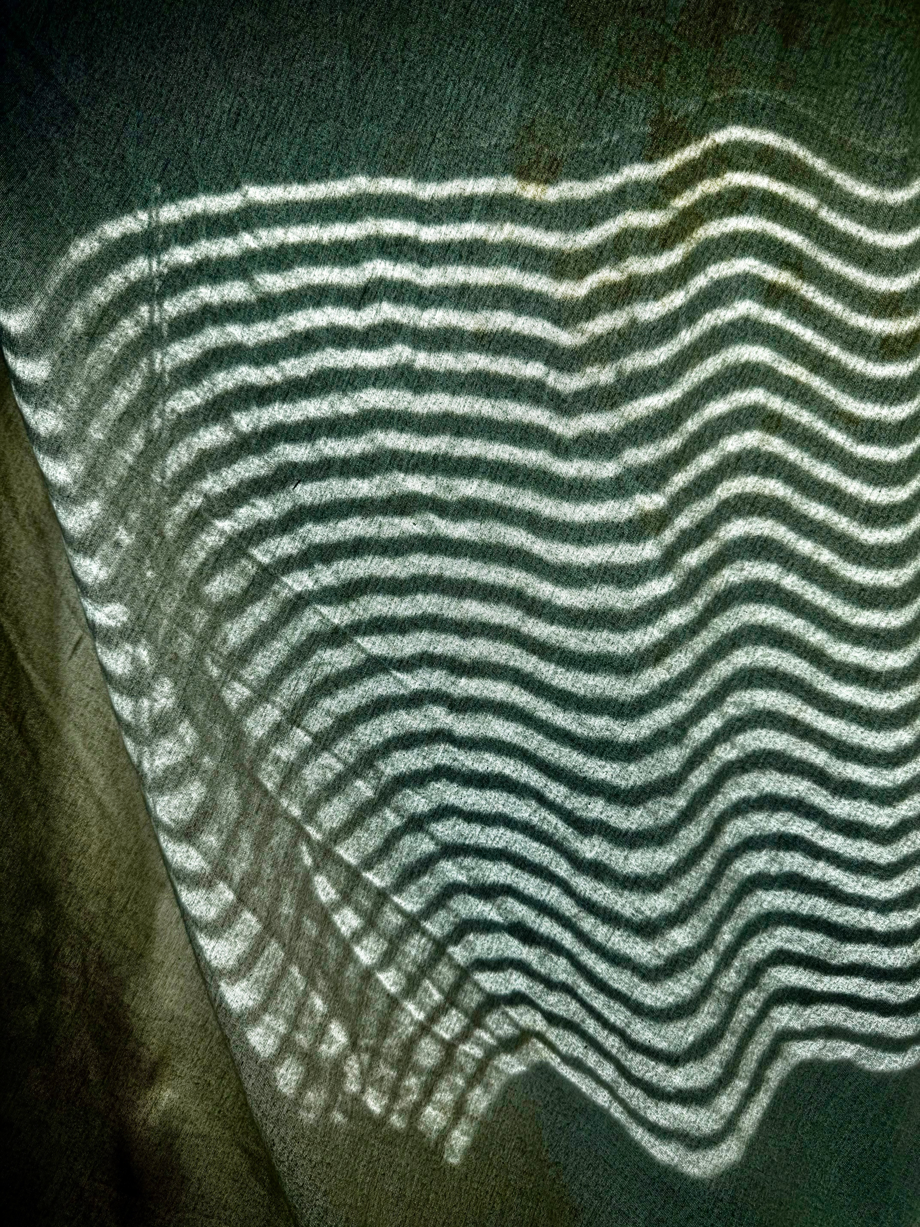 Hard, wavy lines of light on a fitted flannel sheet pulled loosely over a window frame. Bits of scratchy cloth texture catch bits of shadow. Light from slatted blinds curl across the fabric, like the hard shadow lines left by a rake pulled through white sand.