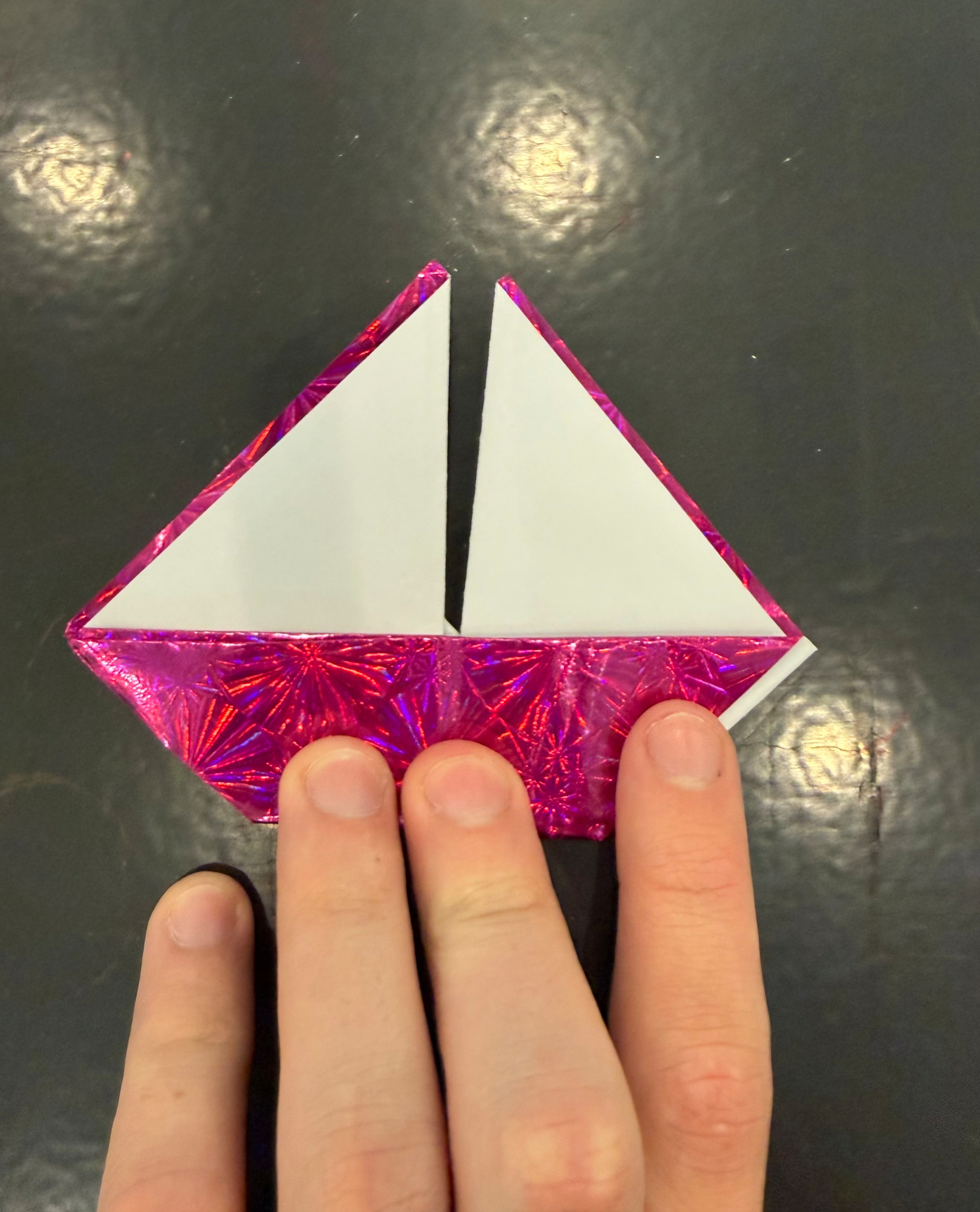 Four fingers touching a paper origami sailboat made with origami paper that has a red glossy coating on one side and nothing on the other, creating very different textures between the front and back. 
