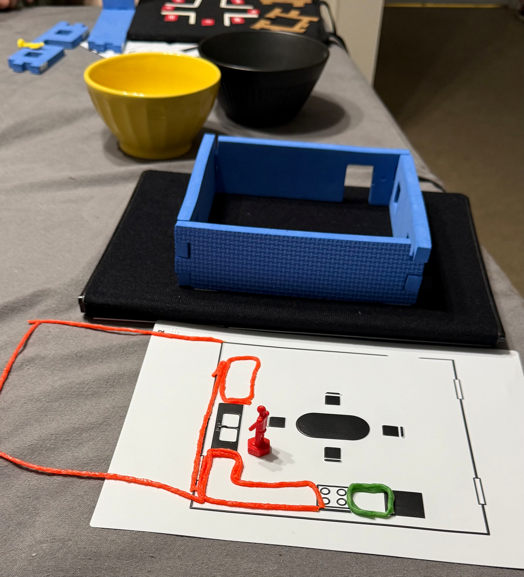A tabletop scene features a small blue model of a room, a floor plan of the same room made with orange wikki stix, a red figurine, and two bowls in the background. The bowls are used to hold 3D version of the 2D shapes in the model. We first read the drawing of the kitchen and then we designed our own additions to the space. 