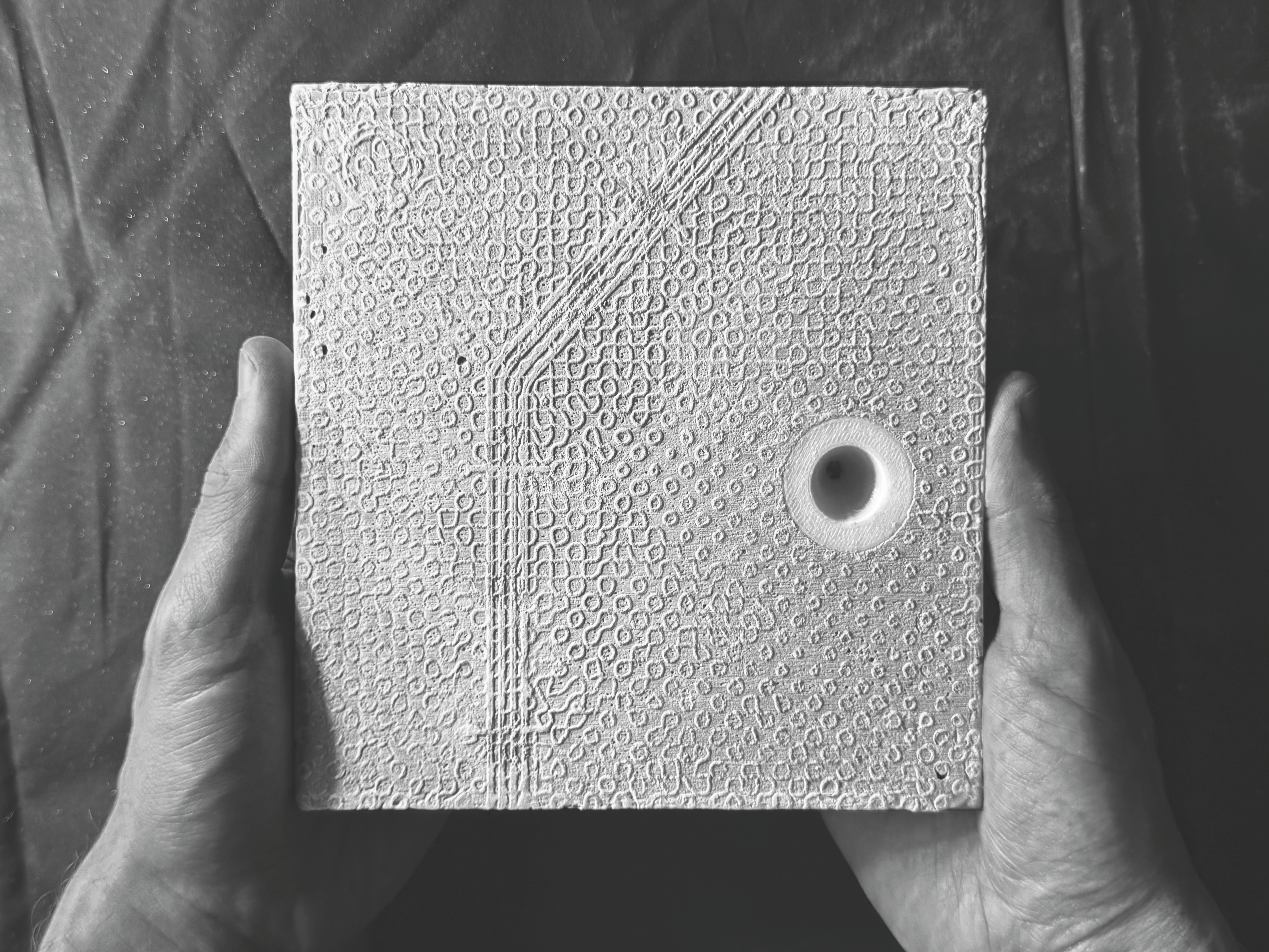 An image of two hands holding a concrete panel with a white hole in it. The panel is cast concrete and the hole is 3D printed, like a trunacated cone inset into the material. There is a rough bitmap texture etched into the concrete but it looks very fragile and irregular, like the image could be wiped away at any moment. And you can see  a bitmap of the city and a subway line. it's a map of a piece of the San Francisco Bay Area, and the map depicts imagined futures / moments of absence and desire for the city. 