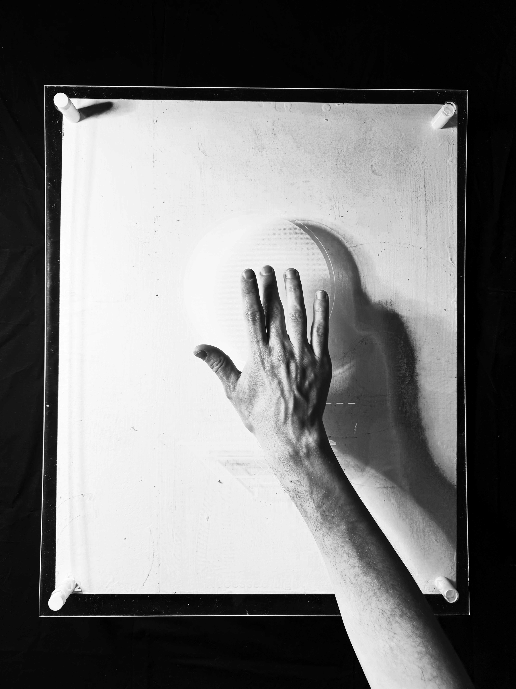 A hand touching a piece of plexiglass mounted on a white surface. We can see the faint impression of a white on white drawing, partly because the hand is casting a shadow through the drawing. The photo is black and white, and the hand has the middle finger pressed down onto the drawing on the surface of the glass. 